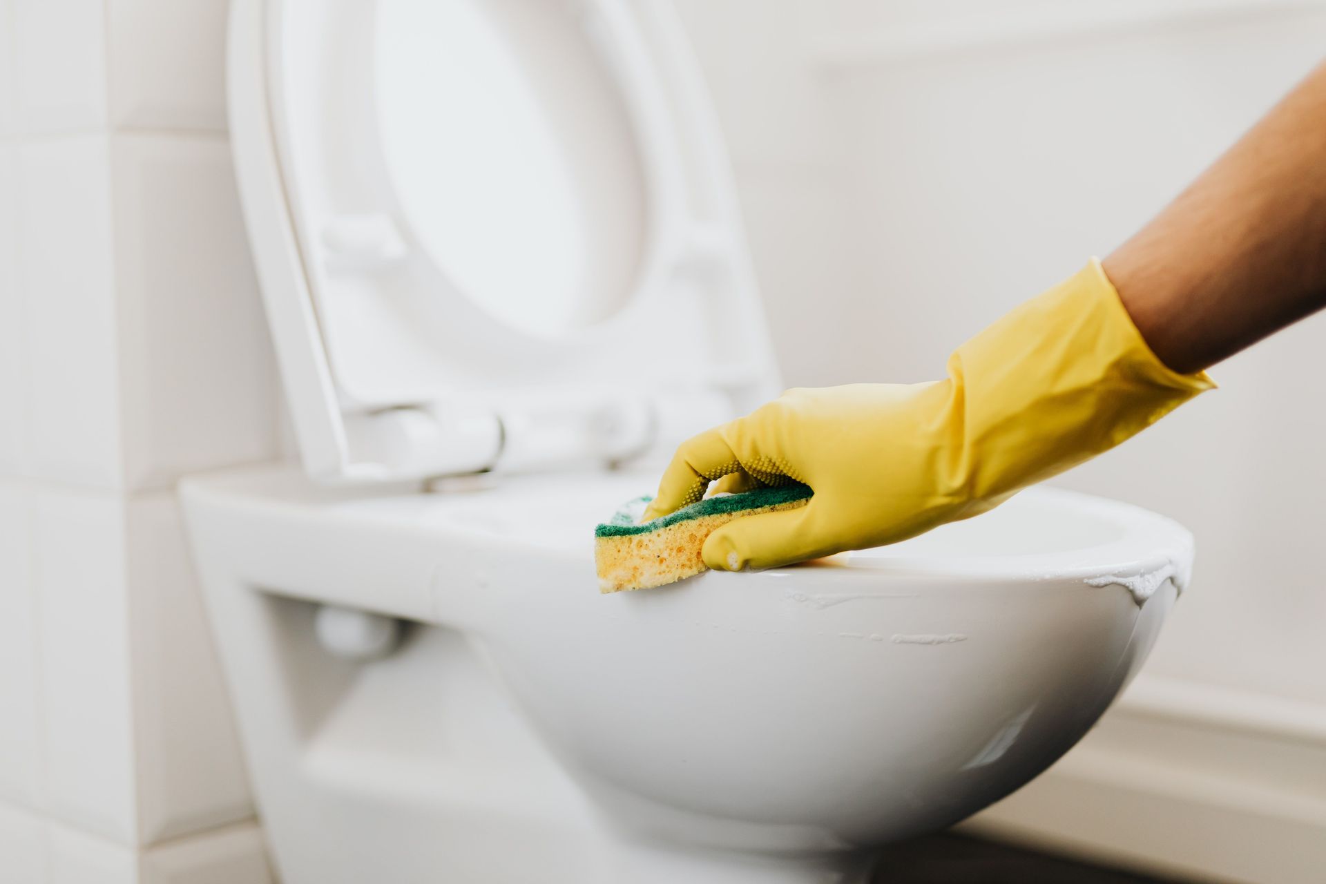 Crop anonymous person in yellow gloves cleaning white toilet rim with detergent and sponge in bathroom